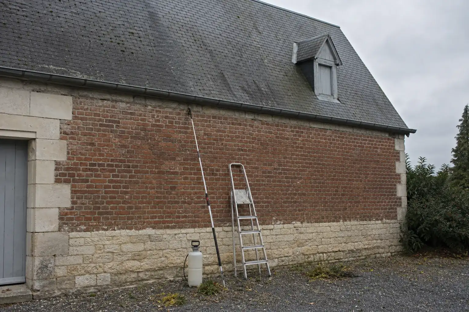 Bidon de produit anti-mousse et pulvérisateur à perche posés au pied d’une maison
