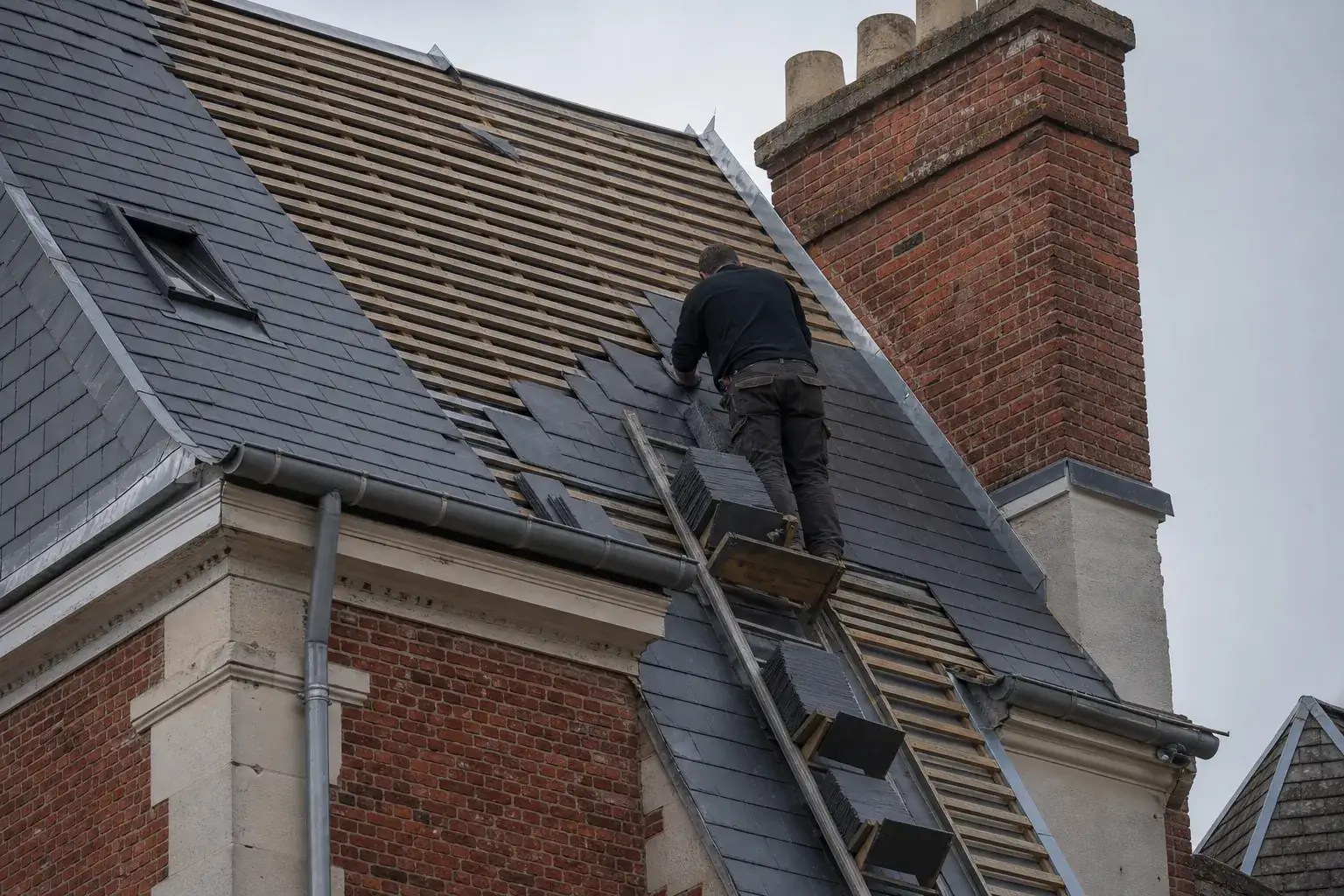 Versant en ardoise en cours de rénovation sur une maison de brique, ardoises neuves posées par rangs depuis l’égout